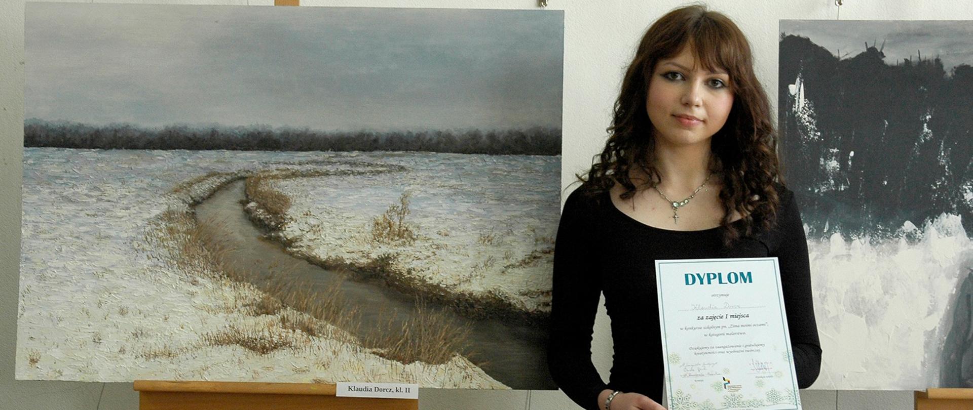 Na zdjęciu z prawej strony uczennica z dyplomem w dłoniach z lewej na sztaludze obraz z motywem krajobrazu zimowego