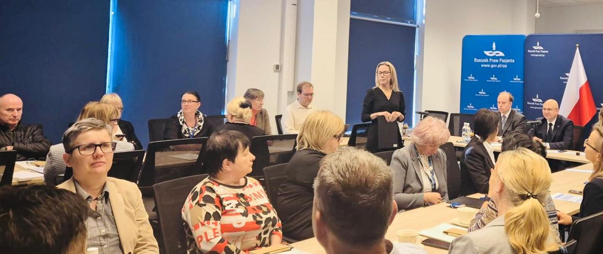 Zdjęcie przedstawia wiele osób siedzących na sali konferencyjnej przy podłużnych stołach, przy stole prezydialnym 3 osoby, w tle ścianka materiałowa z jednym dużym logiem Rzecznika Praw Pacjenta oraz kilkoma małymi, białe loga na granatowym tle. Wszyscy uczestnicy siedzą z wyjątkiem prowadzącej.