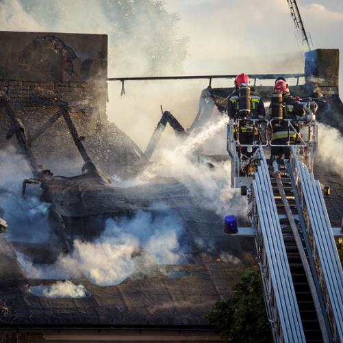 Dwóch strażaków w strojach bojowych na wysuwanej drabinie gasi resztki ognia na dachu domu. Cały dach się zapadł biały dym snuje się poniżej czarnych osmolonych belek dachowych i dwóch murowanych kominów.