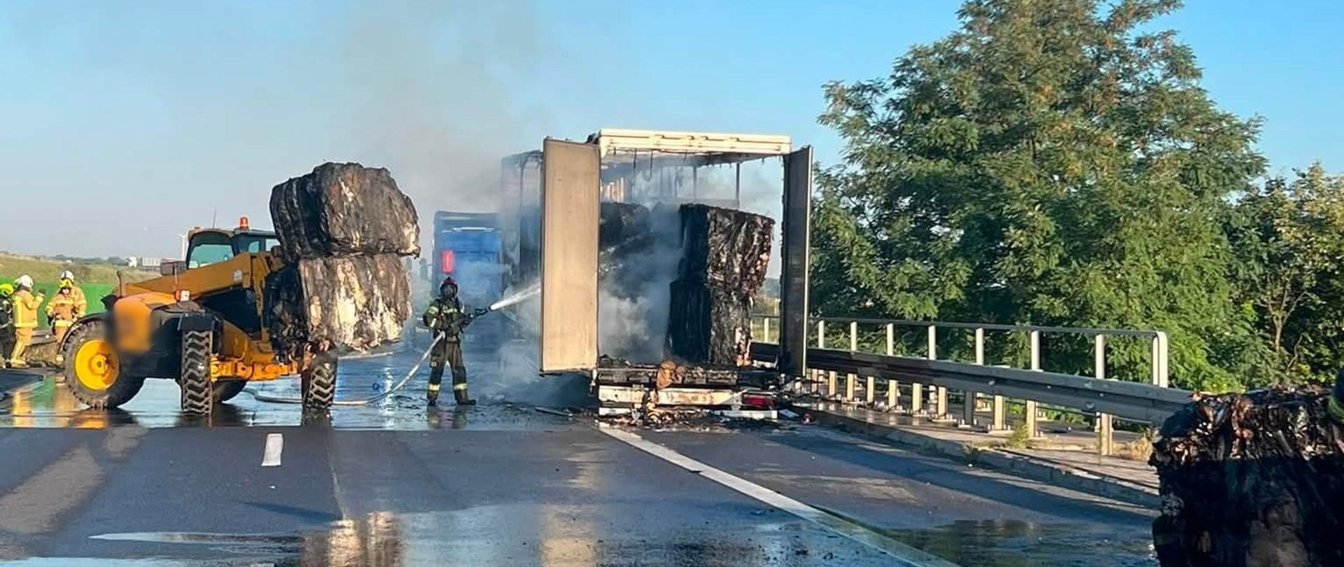 Strażacy gaszą płonący samochód ciężarowy na autostradzie. Z przyczepy unoszą się resztki dymu, a spalone, zwęglone bele ładunku są usuwane za pomocą dużej ładowarki. Kilku strażaków w pełnym umundurowaniu pracuje przy mokrej nawierzchni, kierując strumień wody na tlące się materiały
