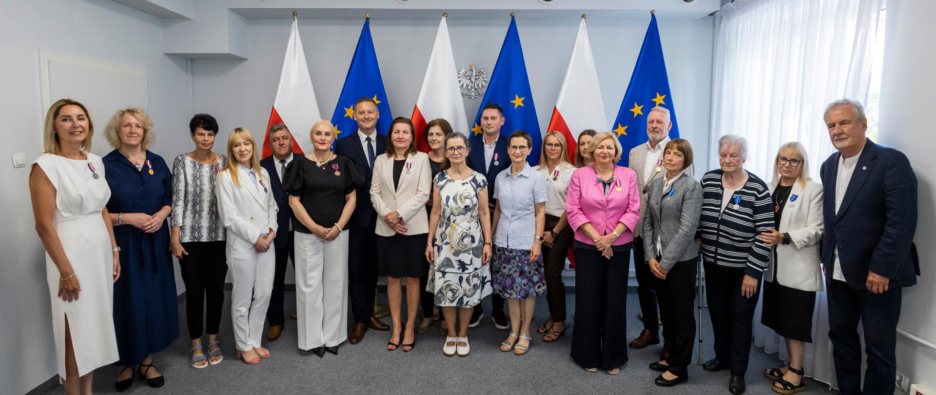 17 odznaczonych osób pozuje w rzędzie do zdjęcia z Wicewojewodą Kujawsko-Pomorskim Michałem Koniuchem na tle flag Polski i Unii Europejskiej oraz godła naszego kraju.