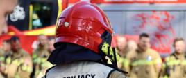 Strażak w czerwonym kasku widziany od tyłu, ubrany w kamizelkę z napisem „KIERUJĄCY DZIAŁANIEM RATOWNICZYM”. W rozmytym tle stoją inni strażacy w ubraniach specjalnych.