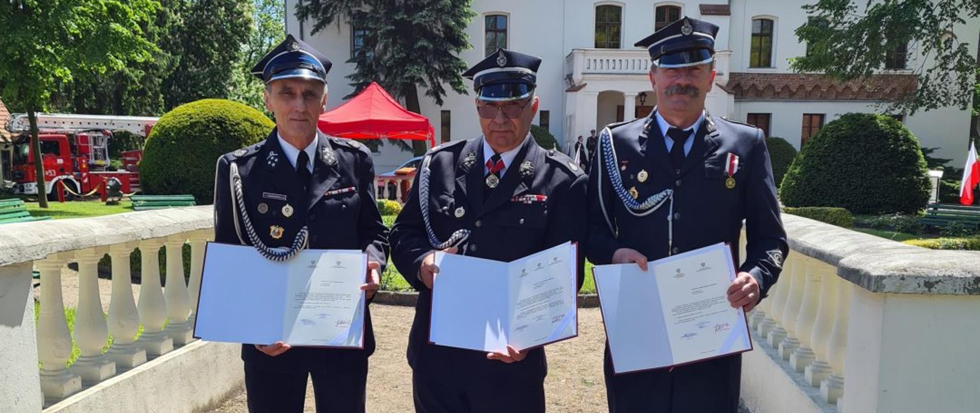 Strażacy w mundurach stoją i trzymają odebrane promesy na zakup nowych samochodów