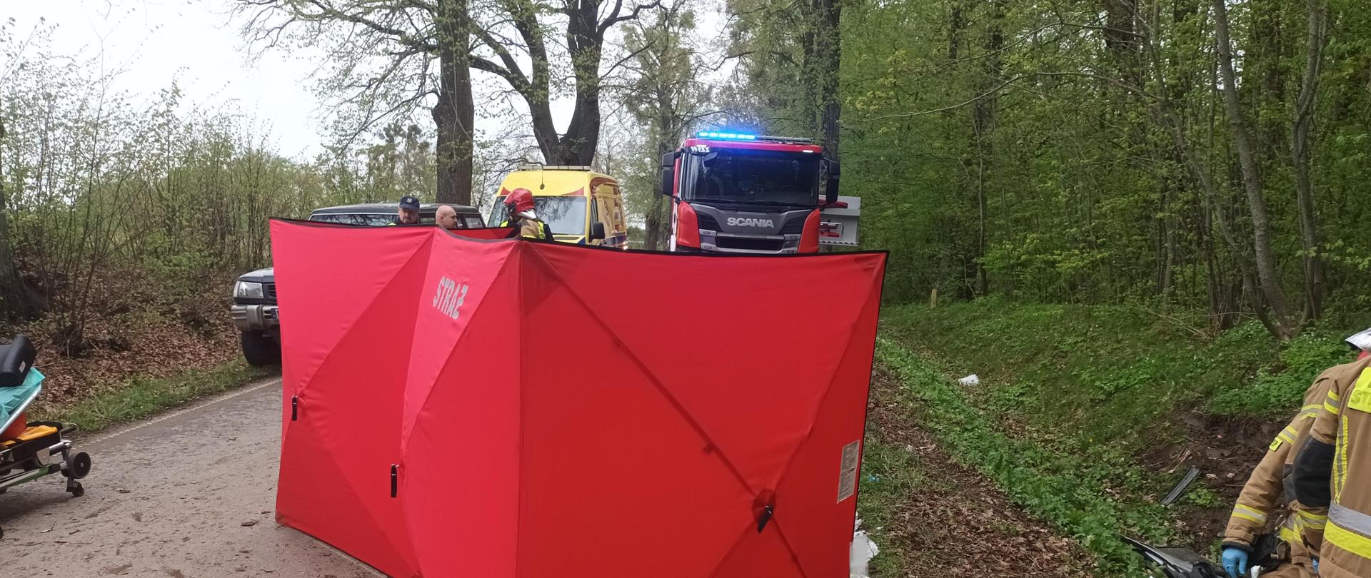 Parawan strażacki w celu zakrycia drastycznych widoków i zachowania prywatności poszkodowanej, w oddali pojazd strażacki i ambulans