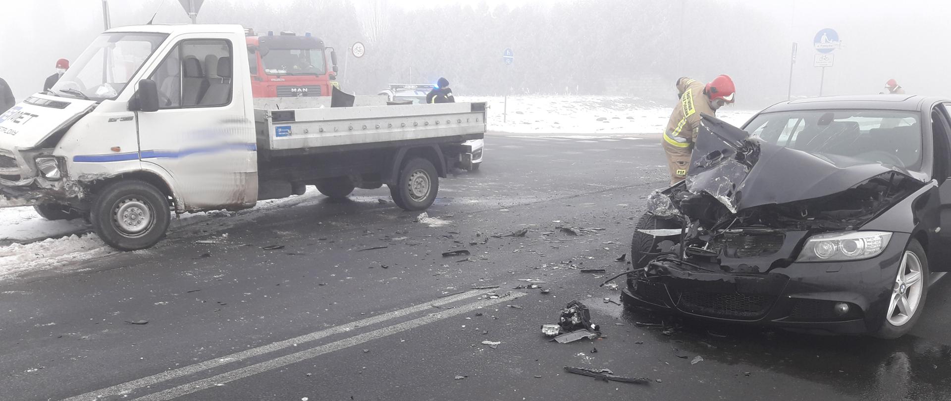 Po lewej stronie rozbity samochód dostawczy, po prawej stronie rozbity samochód osobowy. W tle strażak.