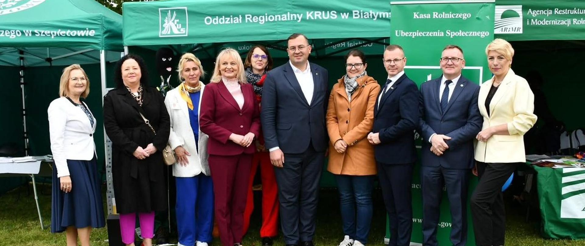 Grupa dziesięciu osób stoi na trawie przed zielonym namiotem z białym napisem „Oddział Regionalny KRUS w Białymstoku”. Za nimi po lewej stronie stoi manekin w zielonym ubraniu roboczym i żółtej kamizelce odblaskowej. Po prawej stronie stoi zielony baner reklamowy z logo KRUS i hasłem „Rolniku, pracuj bezpiecznie”. 