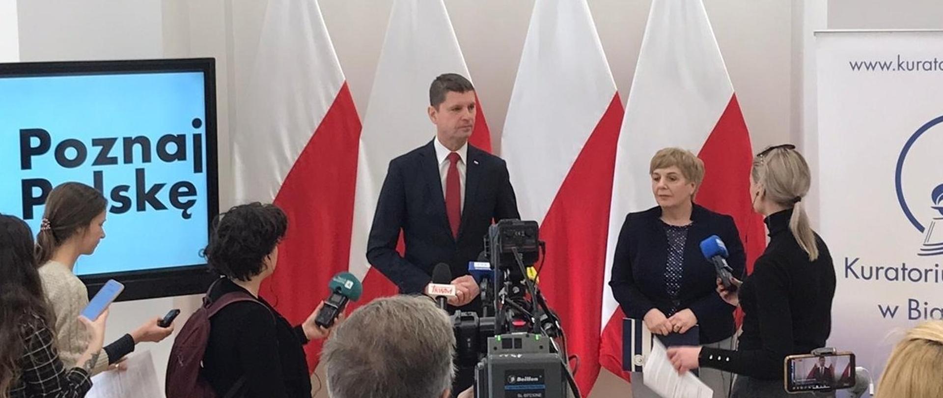 Wiceminister Piontkowski stoi przed dużą kamerą i mówi, obok niego stoi kilka osób, za nimi cztery polskie flagi i duży monitor wyświetlający napis Poznaj Polskę na jasnoniebieskim tle.
