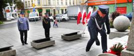 Upamiętnienie rocznicy. Plac przed pomnikiem Żołnierza Polskiego w Rawiczu. Delegacja rawickich służb mundurowych składa wiązankę kwiatów. Funkcjonariusz Służby Więziennej jest pochylony przed pomnikiem, natomiast strażak i policjant stoją w postawie zasadniczej kilka metrów przed pomnikiem. Z prawej strony stojak z flagami państwowymi. W tle budynki, przechodnie i ruch uliczny.