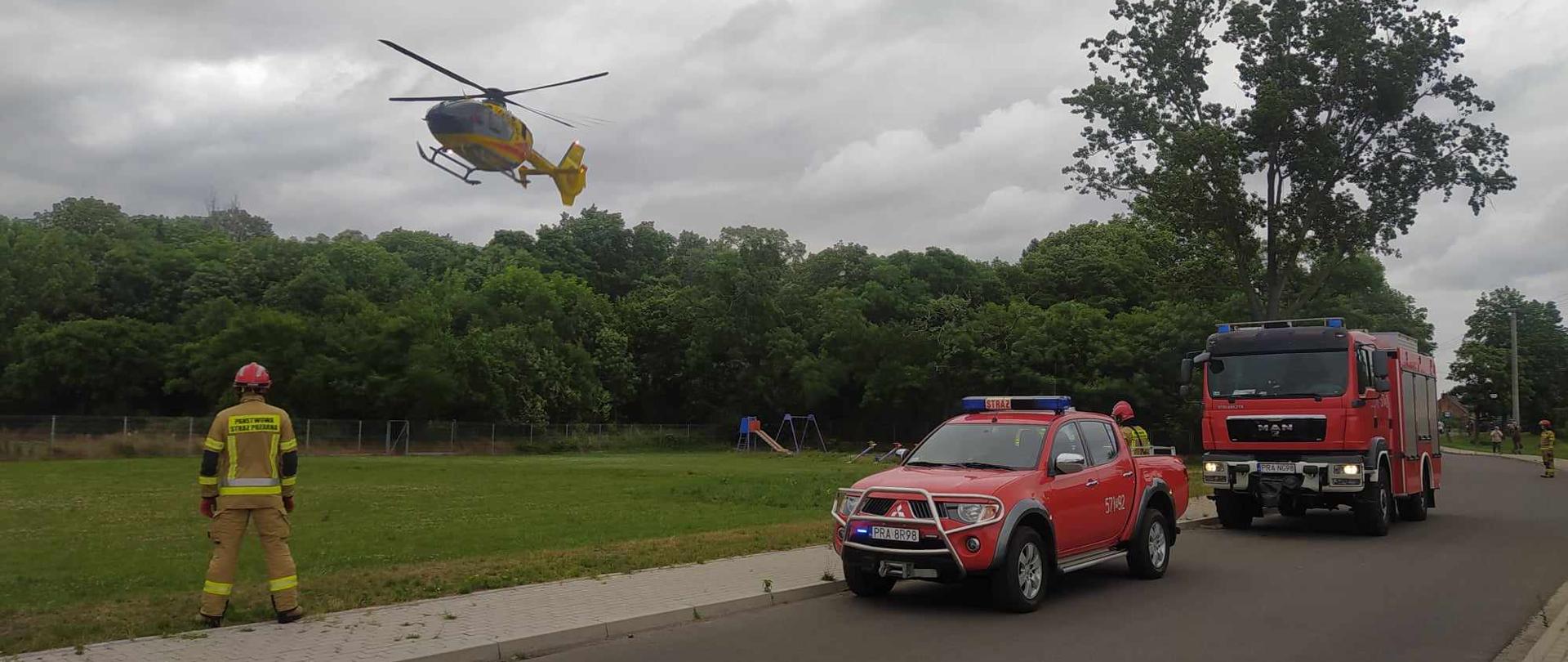 Interwencja LPR. Boisko sportowe w Konarzewie. Do lądowania podchodzi śmigłowiec Lotniczego Pogotowia Ratunkowego. Na drodze obok boiska stoją dwa wozy strażackie. Wokół nich znajduje się 3 strażaków w ubraniach specjalnych koloru piaskowego i w czerwonych hełmach. W tle las i plac zabaw. 