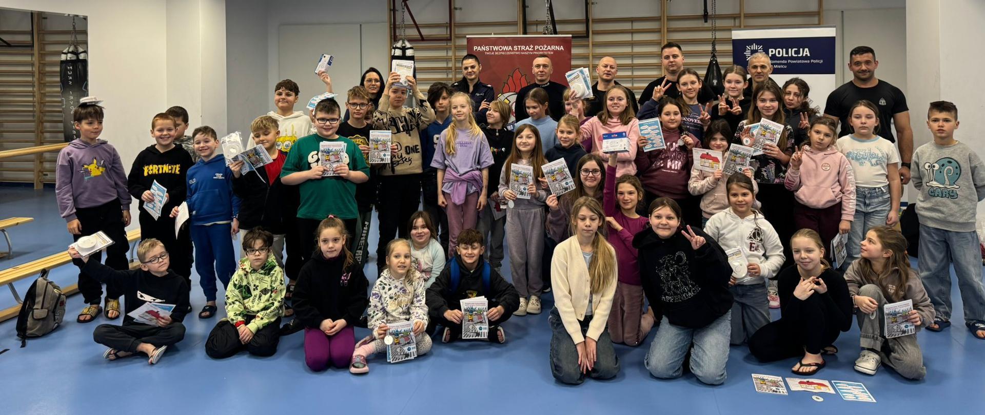Pamiątkowe zdjęcie grupowe dzieci, policjantów i strażaków. Dzieci z uśmiechem prezentują otrzymane upominki: odblaski, broszury informacyjne oraz białe pudełka z czujkami tlenku węgla i dymu.