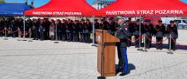 Pomorski komendant wojewódzki Państwowej Straży Pożarnej wita gości, pod namiotami stoją zaproszeni na uroczystość. 