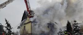 Widoczny pożar budynku mieszkalnego, silne zadymienie, podnośnik hydrauliczny i strażacy pracujący na nim