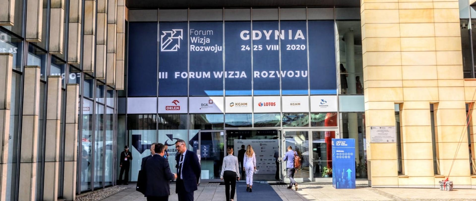 Na zdjęciu wejście do budynku biblioteki Akademii Marynarki Wojennej w Gdyni, gdzie odbyło się forum. Widoczni rozmawiający ludzie. Na budynku informacje nt. wydarzenia - data (24-25 sierpnia), Gdynia, logo forum.