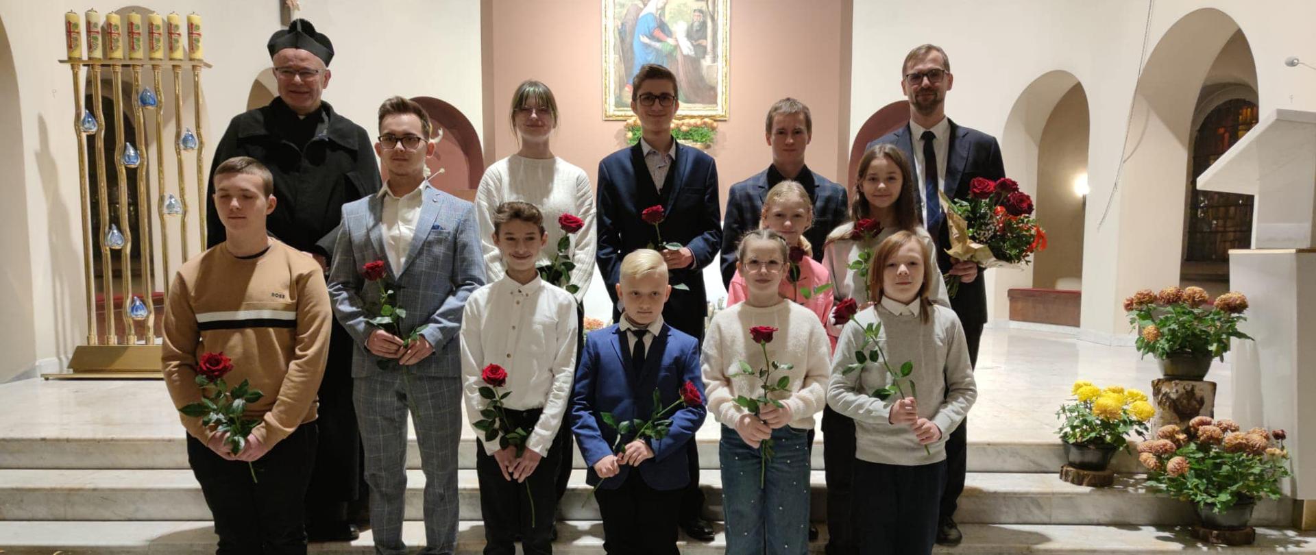 Grupa uczniów stoi przed prezbiterium kościoła, w rękach trzymają kwiaty