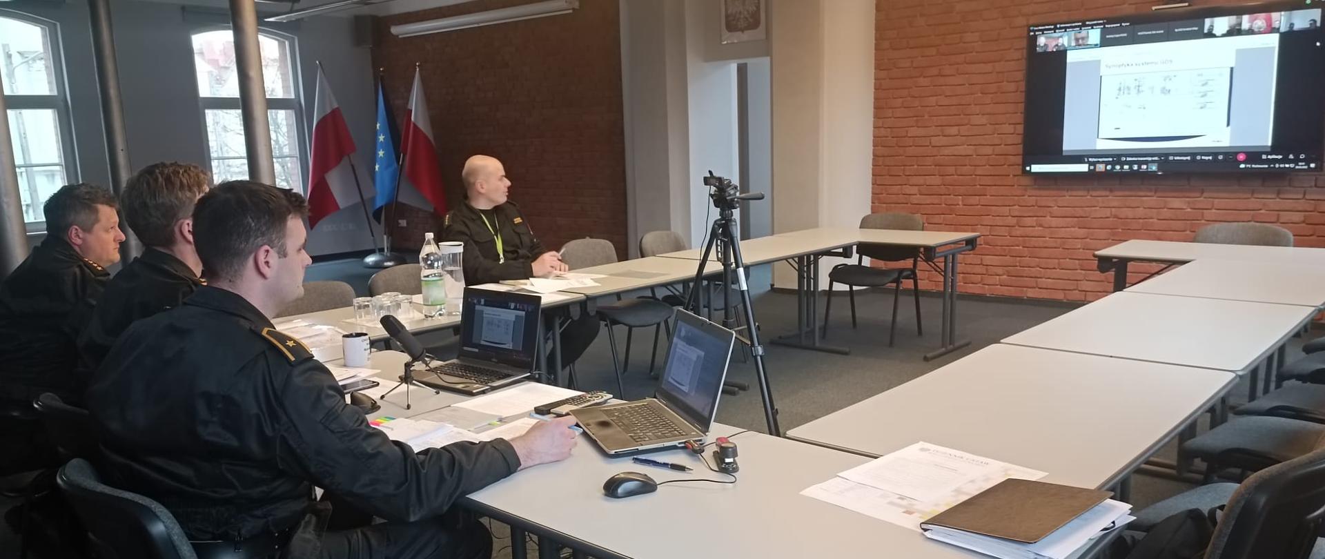 czterech strażaków siedzi przy stole na którym znajdują sie komputery, w tle widać flagownik z flagami biało czerwonymi oraz niebieską z żółtymi gwiazdkami, przed strażakami wisi telewizor