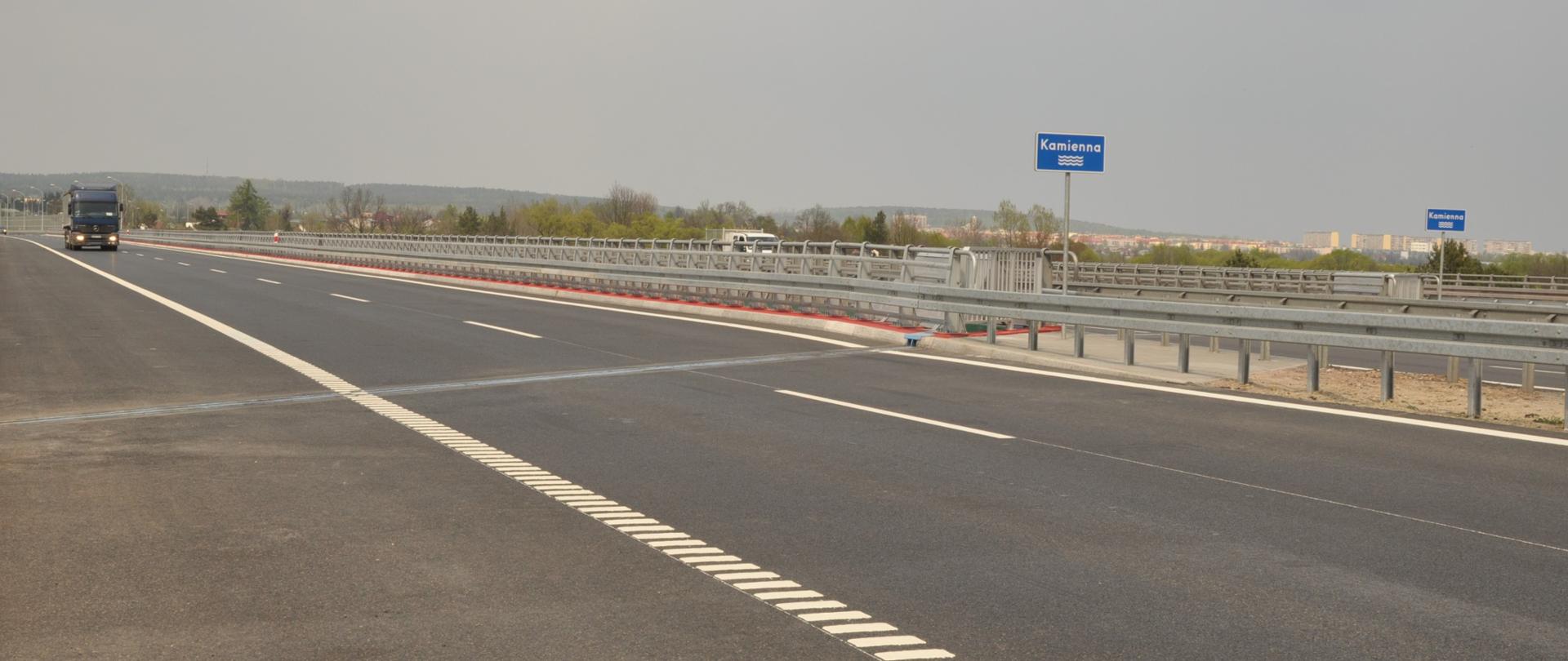 Estakada S7 Skarżysko-Kamienna - na pierwszym planie jedna jezdnia dwujezdniowej estakady, bariery w pasie rozdziału, w oddali jadący samochód. Przy drodze niebieska tablica z nazwą rzeki "Kamienna"