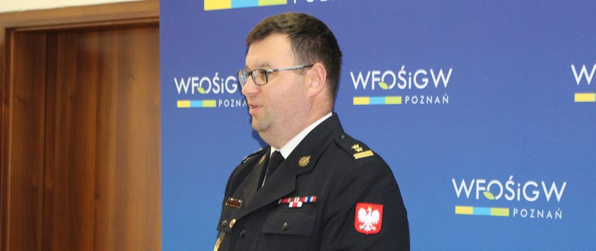 strażak w mundurze wyjściowym stoi przed mównicą, za mężczynzą stoi niebieska ściana z napisami WFOŚIGW POZNAN