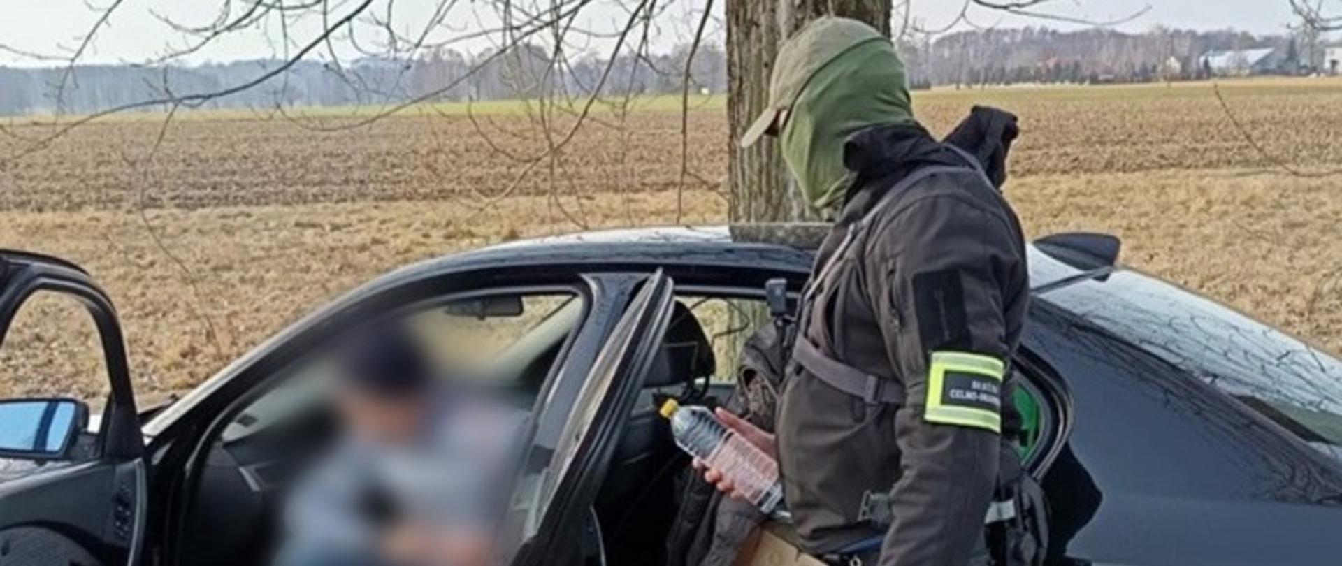 Umundurowany funkcjonariusz trzyma w ręku plastikową butelkę z żółtym korkiem, stojąc przy samochodzie osobowym. 