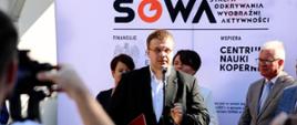 Mężczyzna w garniturze stoi pod ścianą z napisem SOWA i mówi do mikrofonu.