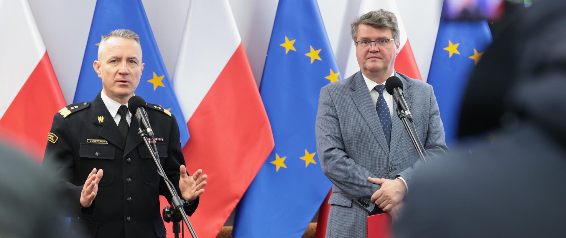 Mężczyzna w strażackim mundurze generalskim oraz drugi w okularach i szarym garniturze stojący przed mikrofonami. Za nim flagi Polski oraz Unii Europejskiej. Na pierwszym planie widoczni dziennikarze biorący udział w konferencji