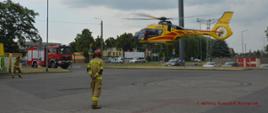 Wspólna akcja strażaków i LPR. Parking jednego z rawickich hipermarketów. W powietrze wzbija się śmigłowiec Lotniczego Pogotowia Ratunkowego. Z lewej strony wóz straży pożarnej. Na parkingu strażacy zabezpieczający operacje startu. W tle drzewa, budynki i samochody.