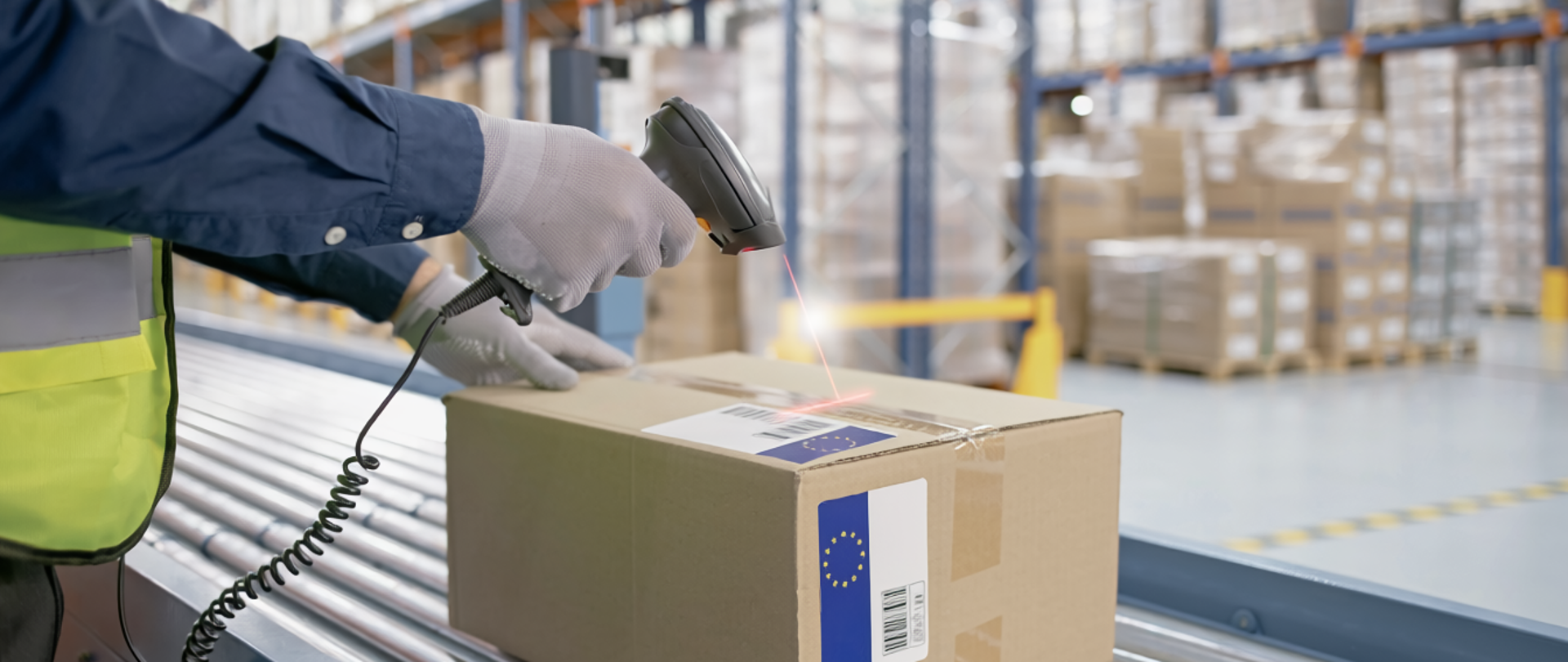 Osoba w kamizelce odblaskowej skanuje kod kreskowy na kartonie, leżącym na taśmie transportowej w magazynie pełnym regałów z zapakowanymi przesyłkami.