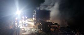 Zdjęcie przedstawia całkowicie spalony ciągnik siodłowy, który stoi na jedni. Pora nocna. Nad samochodem unosi się dym. Na zdjęciu widać również strażaków oraz samochody strażackie. 
