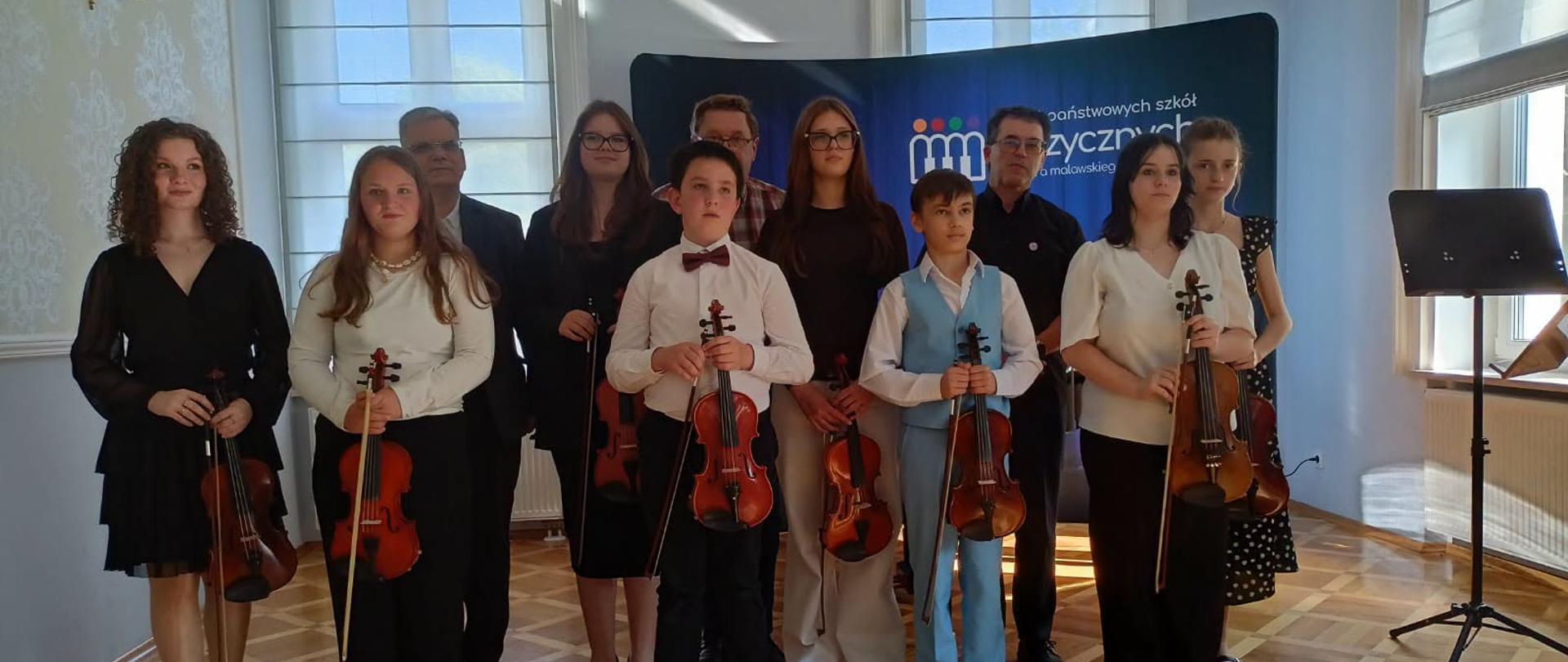 Grupa dzieci, młodzieży i dorosłych pozuje do wspólnego zdjęcia w jasnej sali koncertowej z drewnianą podłogą. Większość uczestników trzyma skrzypce i smyczki, ubrana jest w eleganckie stroje koncertowe w czerni i bieli. W tle znajduje się granatowy baner z logo państwowych szkół muzycznych oraz wysokie okna wpuszczające światło dzienne.