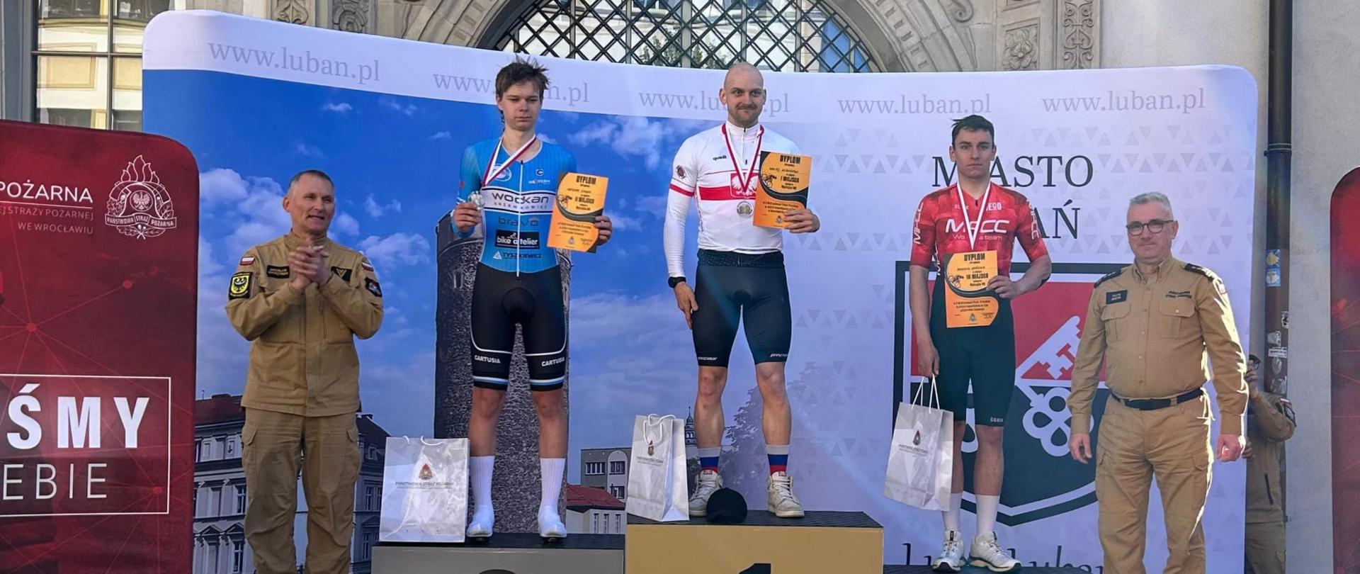 Osoby biorące udział w zawodach stoją w strojach kolarskich na trójstopniowym podium. Na szyjach mają medale, w rękach dyplomy. Po prawej i lewej stronie poza podium stoją funkcjonariusze PSP w żółtych ubraniach służbowych. 