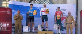 Osoby biorące udział w zawodach stoją w strojach kolarskich na trójstopniowym podium. Na szyjach mają medale, w rękach dyplomy. Po prawej i lewej stronie poza podium stoją funkcjonariusze PSP w żółtych ubraniach służbowych. 