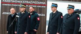 Przed budynkiem Komendy Powiatowej Państwowej Straży Pożarnej w Janowie Lubelskim kilku oficerów PSP w mundurach wyjściowych stoi przy mikrofonie podczas oficjalnego wystąpienia. W tle widoczna jest tablica z nazwą jednostki.