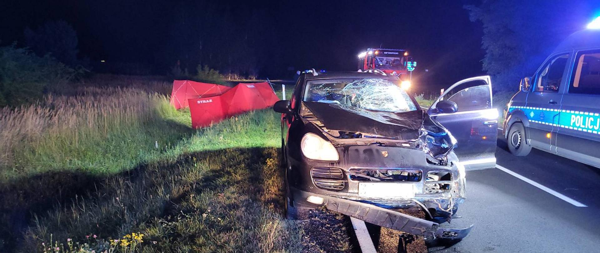 Zdjęcie przedstawia samochód osobowy z uszkodzonym przodem. Zderzak częściowo urwany, zbita przednia szyba, uszkodzony reflektor. Z tyłu auta w przydrożnym rowie rozstawiony parawan strażacki koloru czerwonego. Na zdjęciu widać również samochód strażacki oraz radiowóz. 