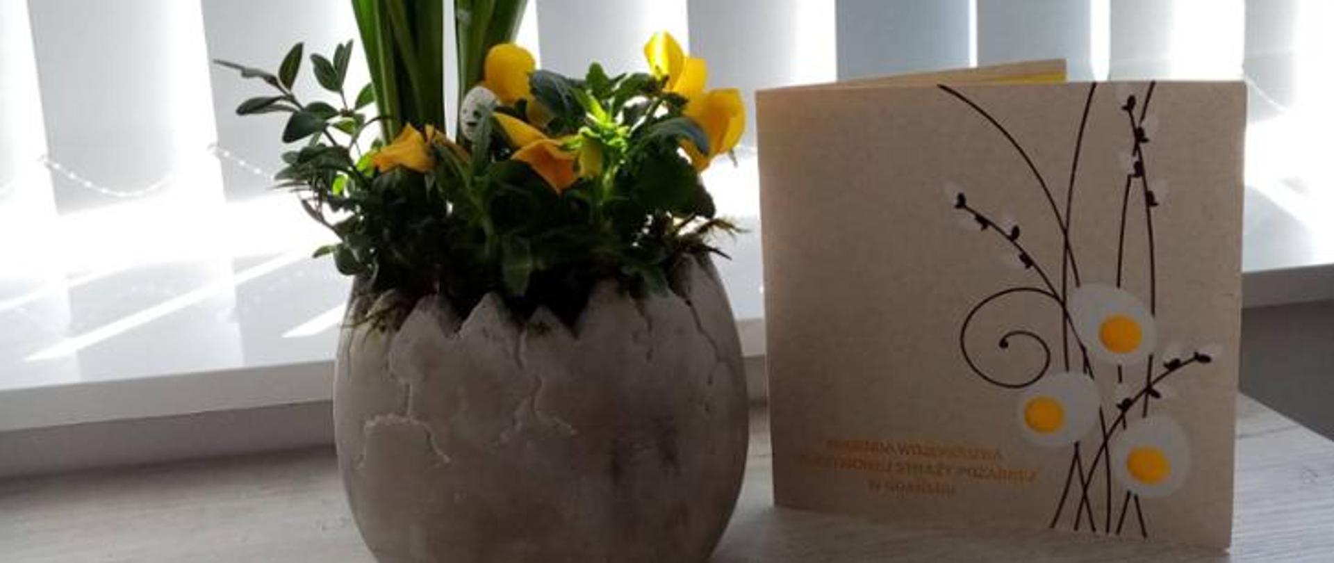 Zdjęcie ukazuje kartkę z życzeniami oraz obok stoi wazon w kształcie jaja a w nim kwiaty.