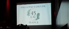 Zdjęcie przedstawia planszę z napisem "PSM I stopnia w Jarocinie - 45 lat tradycji" wyświetlaną przez rzutnik na ekranie podczas uroczystości obchodów jubileuszu szkoły.