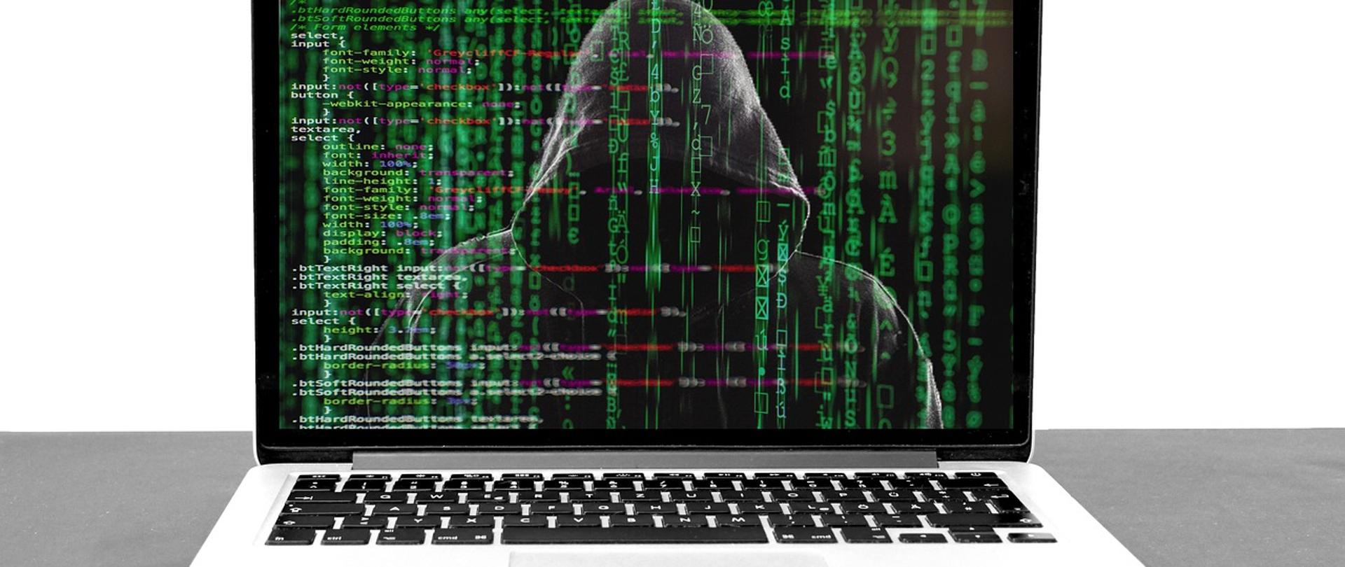 Laptop, na ekranie zdjęcie hakera w czarnej bluzie z kapturem, na nim ciąg zielony liter i znaków