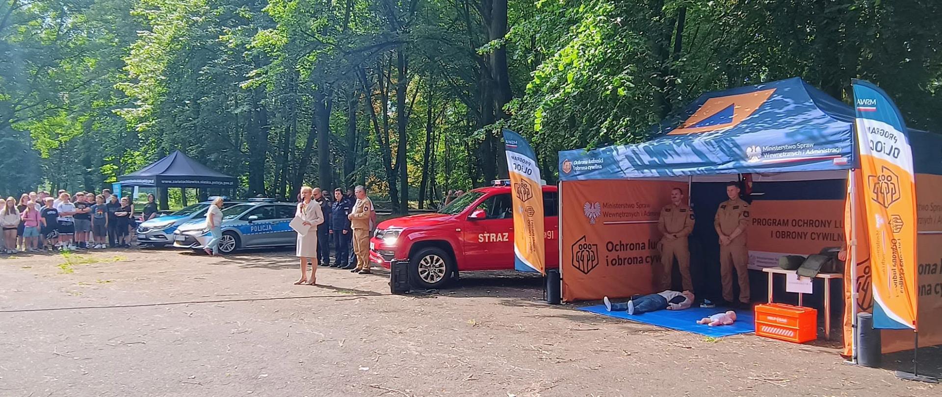 Na zdjęciu zaprezentowano oficjalną część plenerowego wydarzenia, która odbywa się na placu w otoczeniu gęstych drzew. W centralnej części kadru stoi kobieta w jasnej garsonce, trzymająca w rękach dokumenty, która przemawia do zgromadzonych. Obok niej, w równym szeregu, stoją funkcjonariusze policji w ciemnych mundurach oraz strażacy w piaskowych strojach służbowych. Za nimi zaparkowane są pojazdy operacyjne: dwa oznakowane radiowozy policyjne oraz czerwony samochód terenowy Państwowej Straży Pożarnej.
Po prawej stronie znajduje się duży, zadaszony namiot promocyjny z napisem „Program Ochrony Ludności i Obrony Cywilnej” oraz logiem Ministerstwa Spraw Wewnętrznych i Administracji. Pod namiotem stoją dwaj strażacy w piaskowych mundurach, a przed nimi, na niebieskiej macie rozłożonej na ziemi, umieszczono manekiny służące do nauki pierwszej pomocy. W głębi placu, po lewej stronie, widoczna jest grupa młodzieży oraz kolejny, ciemny namiot rozstawiony wśród drzew.