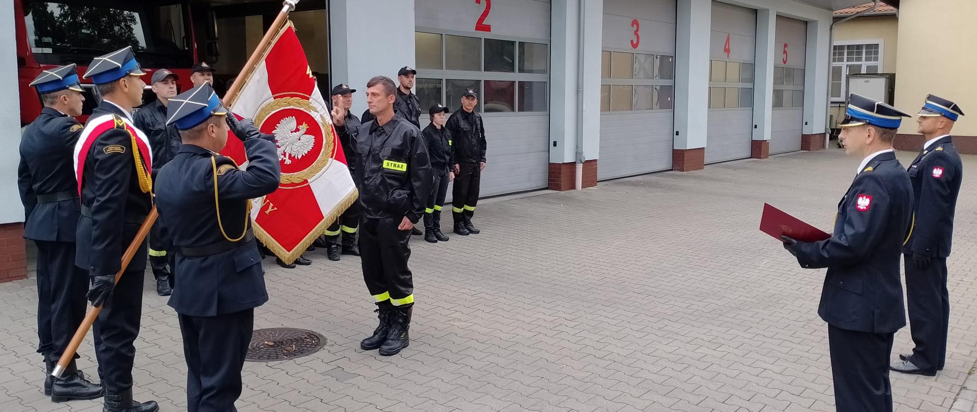 Po prawej stronie prowadzący uroczystość i zastępca komendanta, po lewej poczet sztandarowy z opuszczonym do ślubowania sztandarem i stojący przed nim ślubujący strażak, w tle budynek garażowy.