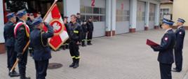 Po prawej stronie prowadzący uroczystość i zastępca komendanta, po lewej poczet sztandarowy z opuszczonym do ślubowania sztandarem i stojący przed nim ślubujący strażak, w tle budynek garażowy.