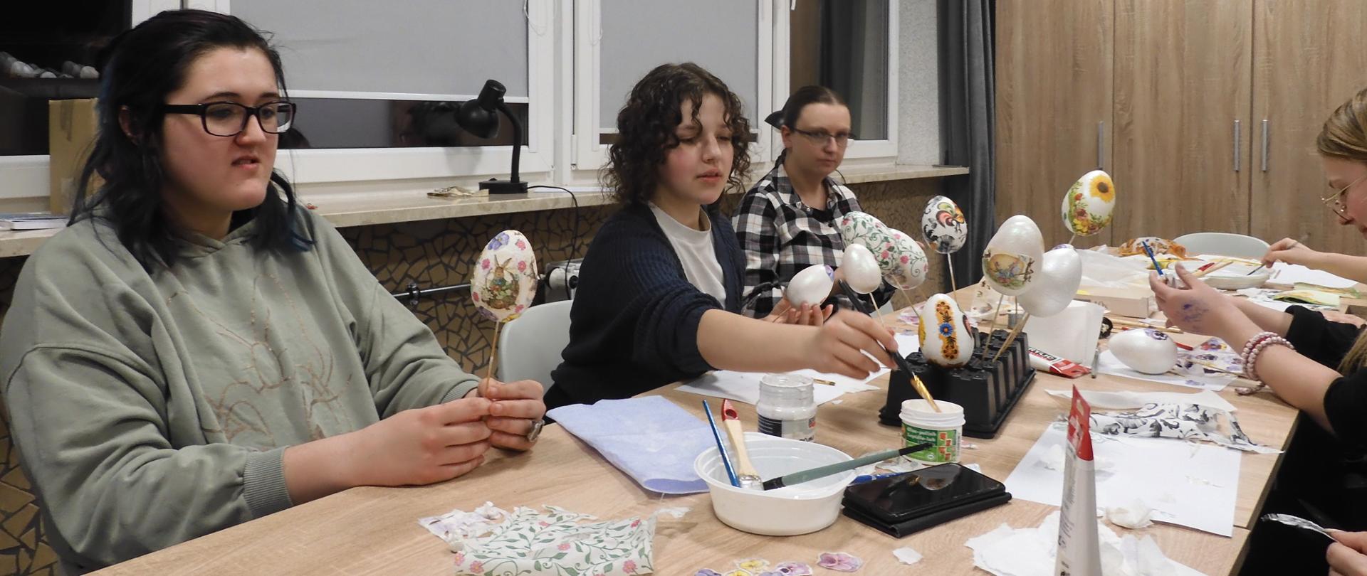 zdjecie w swietlicy, przy podłuznym stole siedzi grupa młodych ludzi, którzy ozdabiają pisanki. Na stole liczne przybory plastyczne czyli kartki, farby , pędzelki, talerzyki plastikowe i styropianowe pisanki.