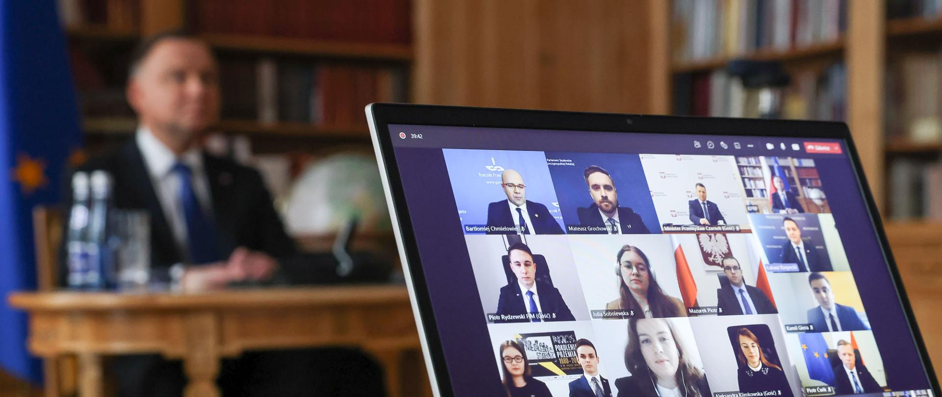Na zdjęciu prezydent Duda podczas wideokonferencji, przed nim laptop na którym widać uczestników.