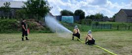 Dwóch młodych strażaków w strojach OSP (żółte koszulki i czarne spodnie z szelkami) klęczy na trawie podczas zawodów sportowo-pożarniczych, trzymając wąż strażacki i kierując silny strumień wody w stronę celu. W tle widać sędziego w czarnym stroju z czerwonym zeszytem, a także wiejską scenerię z domami, ogrodzeniem i drzewami. Pogoda jest słoneczna, niebo z lekkim zachmurzeniem.