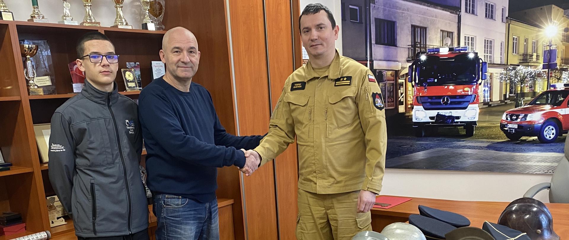 Komendant Powiatowy PSP z Panem Robertem Łuczakiem na tle przekazywanego umundurowania