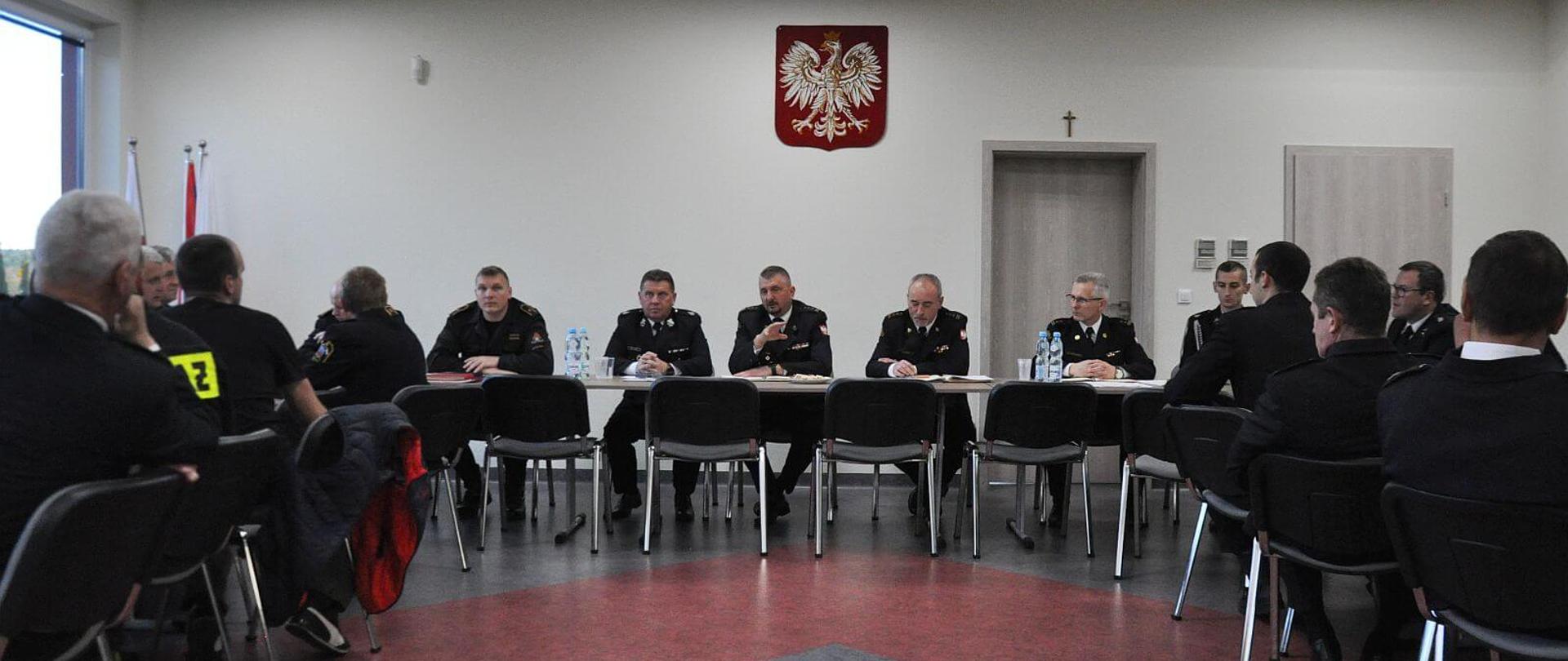 strażacy siedzący w trakcie spotkanie przy stołach w sali konferencyjnej, na ścianie sali widoczne jest godło Rzeczpospolitej Polskie