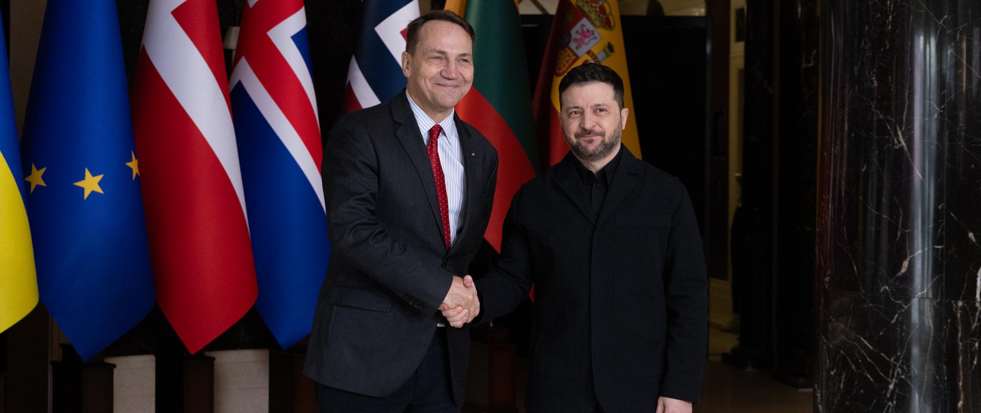 Na zdjęciu jest dwóch mężczyzn w ciemnych garniturach (Wicepremier, Minister Spraw Zagranicznych RP Radosław Sikorski oraz Prezydent Ukrainy Wołodymyr Zełenski). Podają sobie dłonie. Za nimi są flagi państw. 