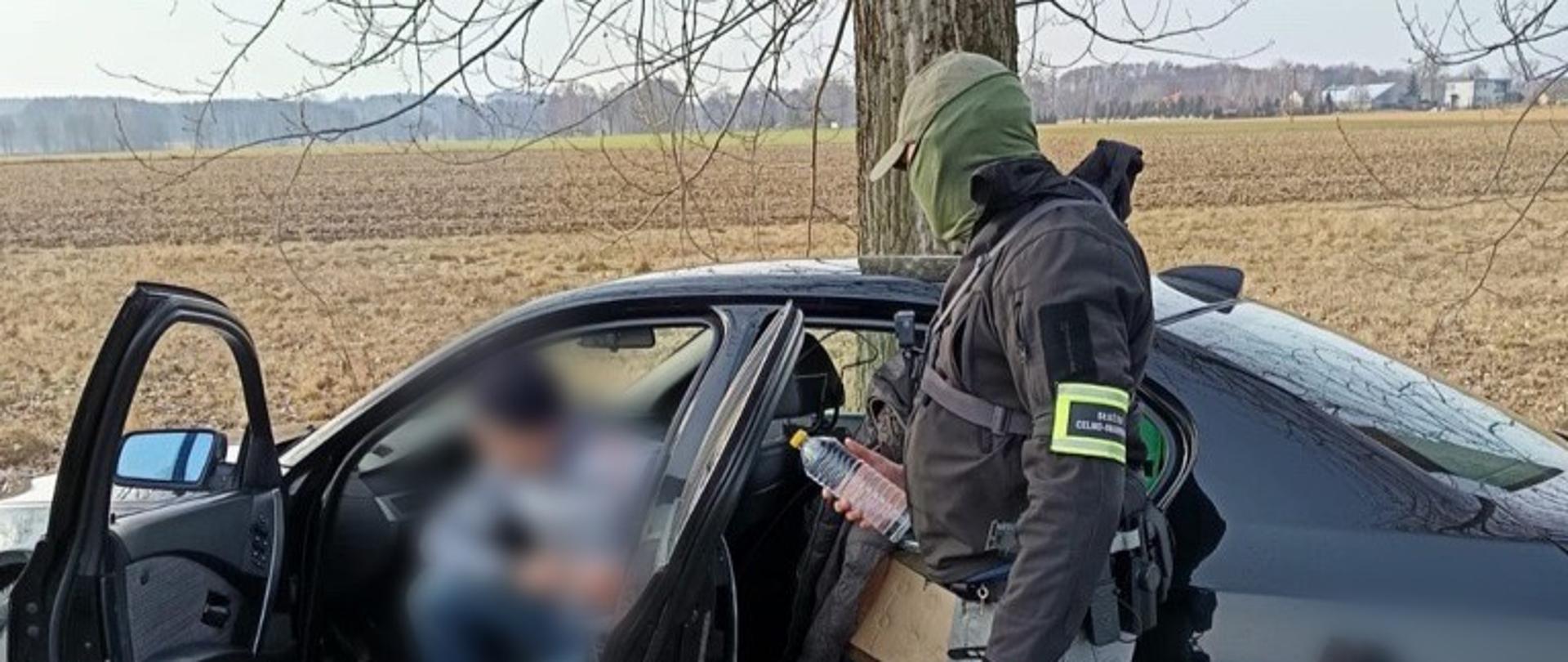 Funkcjonariusz przy otwartych drzwiach czarnego samochodu ogląda plastikową butelkę trzymaną w dłoni.