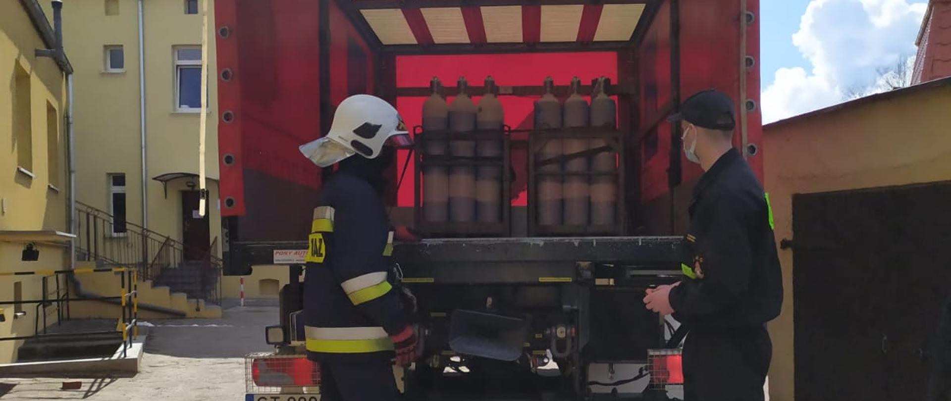 Na zdjęciu widoczny od tyłu czerwono-biały ciężarowy samochód strażacki stojący na utwardzonym terenie. Za pojazdem stoją dwaj strażacy. Jeden w mundurze koszarowym, drugi w mundurze bojowym i hełmie. Na otwartym przedziale ładunkowym znajdują się 2 metalowe kosze zawierające pakiet białych butli stalowych. W tle błękitne, częściowo zachmurzone niebo oraz budynki w kolorze kremowym.