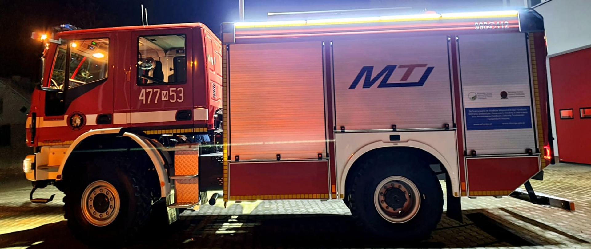 Na zdjęciu przed strażnicą OSP Janówek Góra nowy wóz bojowy bokiem. Widać numer operacyjny 477M53. Zdjęcie wieczorem pojazd z zapalonymi światłami ledowymi oraz masztem oświetleniowym. 