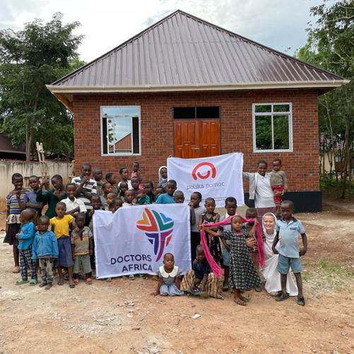 Grupa kilkudziesięciu osób (głównie dzieci) pozuje do zdjęcia przed nowowybudowanym parterowym budynkiem. Trzymają przed sobą banery z logotypami Doctors Africa i polska pomoc.