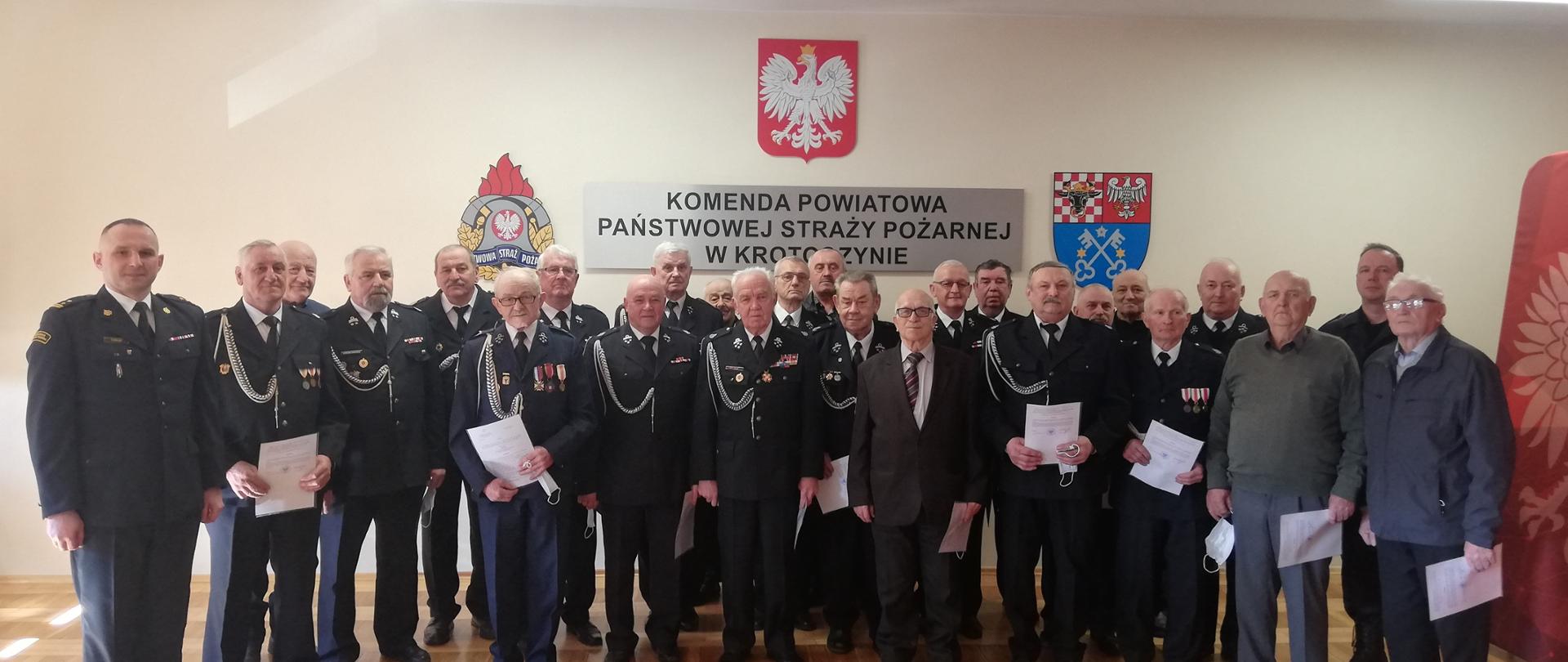 Zdjęcie przedstawia strażaków w mundurach Ochotniczej Straży Pożarnej i Państwowej Straży Pożarnej na tle ściany z logotypem PSP i powiatu krotoszyńskiego oraz nazwą jednostki PSP. Strażacy w ręku trzymają decyzję o przyznanym świadczeniu ratowniczym.
