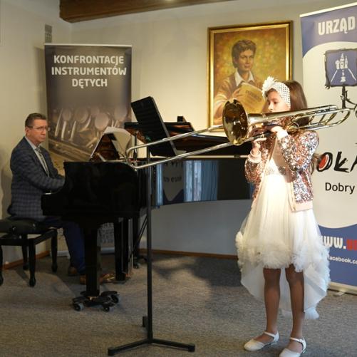Uczennica gra na puzonie stojąc przed fortepianem, mężczyzna gra na fortepianie, w tle banery Konfrontacje Instrumentów Dętych oraz Urząd Miejski w Oławie.